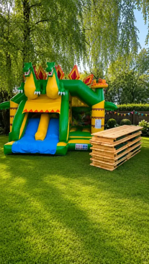 Ein großes, farbenfrohes Bouncy Castle mit Rutsche steht im Garten neben einem Stapel Holzbrettern.